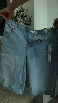 Kädet pitävät avoinna vaaleansinisiä farkkushortsit, joissa on näkyvä vetoketju ja koko-etiketit sisätiloissa vihreän ruukkukasvin lähellä.