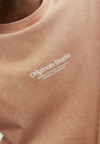 Šviesiai rudas džemperis su apvaliu iškirpimu, švelniu tekstūru, ir balta spauda su užrašu „Originals Studio“ bei prekių ženklu.
