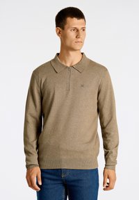 Tröja gjord av stickad tyg, med en dragkedja i kragen i ljusbrun färg, långärmad och med en åtsittande fåll. Diskret logotypdetalj på bröstet.