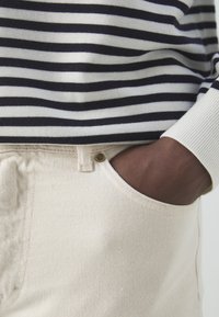 Suéter de punto a rayas en azul marino y blanco, combinado con pantalones de mezclilla claros. Primer plano de una mano en el bolsillo, mostrando la tela texturizada y las costuras.