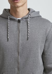 Sivi dukserica sa zatvaračem i vezicama, s glatkom teksturom, prednjim zatvaračem i standardnim krojem. Ugodan i praktičan dizajn.