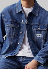 Džínová bunda v středně modré barvě s prsními kapsami, stříbrnými knoflíky a nášivkou Calvin Klein Jeans. Nosit se na bílem tričku.