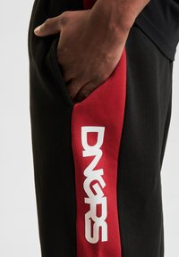 Black joggers with a red side panel featuring white "DNGRS" text, made of soft fabric, with a pocket on the side for convenience.