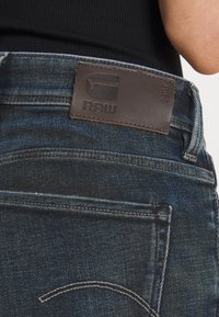 Mörkblå denimjeans med en brun läderbyte på midjan präglad med "RAW," med standardstygn och detaljerade bakfickor.