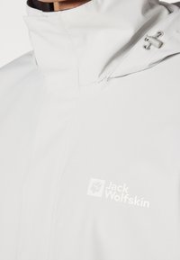 Gros plan d'une veste imperméable gris clair avec un col montant et une capuche ajustable, avec le logo blanc Jack Wolfskin sur la poitrine.