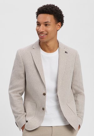 Lächelnder Mann, der einen hellbraunen karierten Blazer über einem weißen Hemd trägt, die Hände in den Taschen, schaut leicht seitlich.