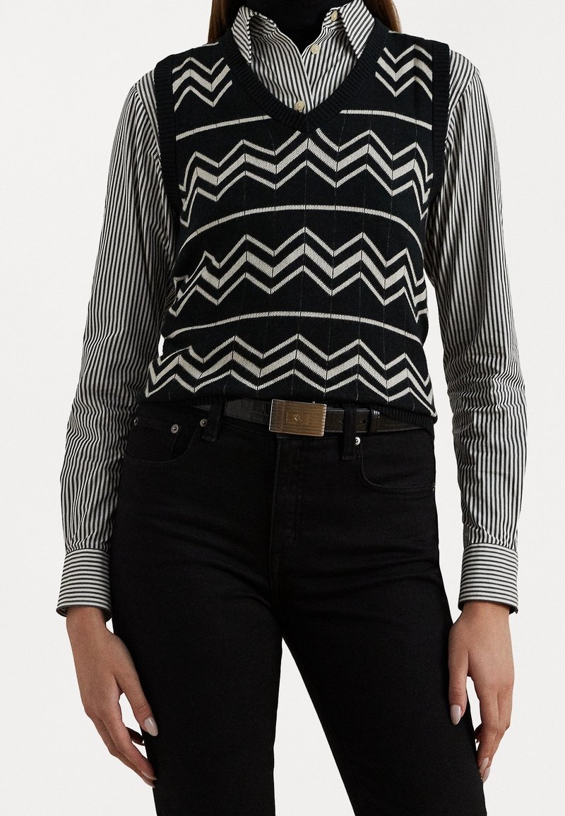 Pull sans manches à motif chevron noir et blanc porté sur une chemise à boutons rayée noir et blanc, associé à un pantalon noir et une ceinture.