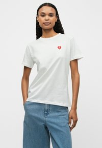 Biała bawełniana koszulka z krótkim rękawem, z małym czerwonym logo serca na lewej piersi. Gładka tekstura i klasyczny krój.