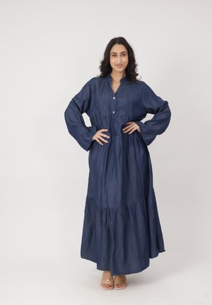 Donna in piedi con le mani sui fianchi, indossa un abito lungo e ampio di colore blu navy con bottoni e maniche larghe, sullo sfondo semplice.