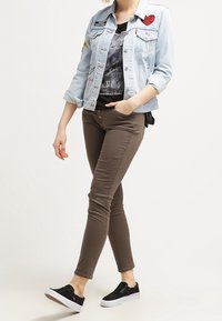 Helles blaues Jeansjacke mit Aufnähern, schwarzes Grafik-T-Shirt, braune enge Hose und schwarze Slipper mit strukturiertem Obermaterial.