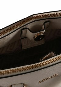 Interieur van een crèmekleurige leren handtas met gouden ritssluiting, voorzien van een zwarte rand, drukknoopsluiting en een patroonvoering. Logo zichtbaar aan de zijkant.