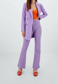 Femme portant un blazer lilas et un pantalon assorti, un top court orange et des sandales à talons rouges, posant avec une main sur la hanche.