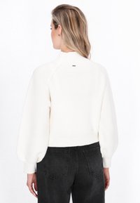 Cremefarvet ribstrikket sweater med høj krave, pufærmer og en lille logo-detajle på bagsiden, parret med mørke jeans.