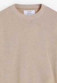 Suéter de punto beige con escote redondo. Textura suave y cuello acanalado. La etiqueta incluye el nombre de la marca y las instrucciones de cuidado.