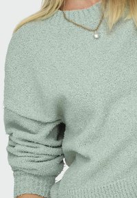 Pull en tricot vert clair avec un tissu texturé et confortable. Doté d'un col rond et de manches légèrement surdimensionnées. Agrémenté d'un collier en chaîne dorée.