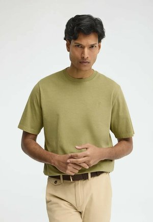 Man die een olijfgroene T-shirt met korte mouwen en beige broek draagt, staand tegen een effen lichtgrijze achtergrond, handen voor zich geklemd.
