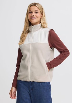 Femme souriante aux longs cheveux blonds portant un gilet polaire beige zippé sur une chemise bordeaux à manches longues et un jean bleu, une main dans la poche.