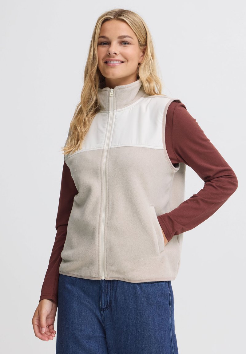 Femme souriante aux longs cheveux blonds portant un gilet polaire beige zippé sur une chemise bordeaux à manches longues et un jean bleu, une main dans la poche.