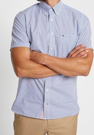 Man met gekruiste armen, draagt een blauw-wit dun gestreept overhemd met korte mouwen en een beige broek tegen een effen achtergrond.