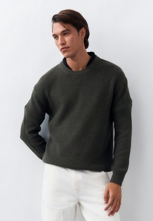 Νέος άνδρας με σκούρα καστανά μαλλιά, φορώντας σκούνο πράσινο πλεκτό πουλόβερ πάνω από μαύρο πουκάμισο και λευκό παντελόνι, στέκεται μπροστά σε απλό ανοιχτό φόντο.