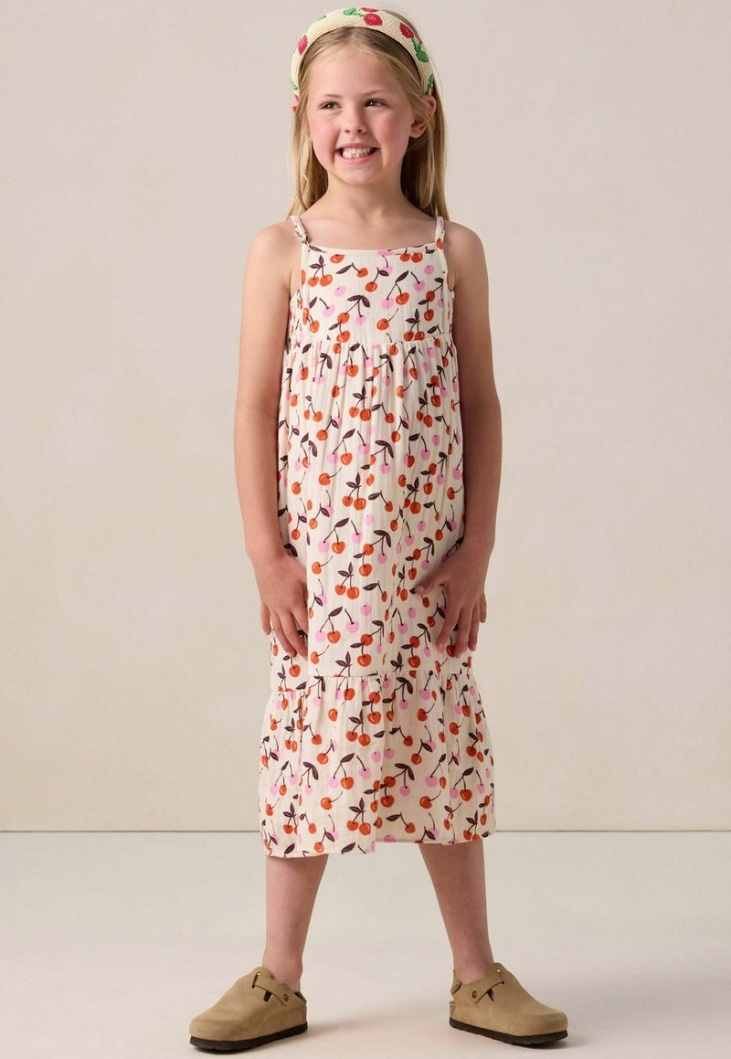 Weißes Kleid mit Kirschmuster, gerafftem Oberteil, gestuftem Rock und dünnen Trägern, kombiniert mit hellbraunen Clogs und einem passenden Stirnband.