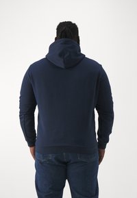 Sudadera azul marino hecha de tela suave, con una gran capucha, mangas largas y puños y dobladillo acanalados. La vista posterior muestra un corte relajado.