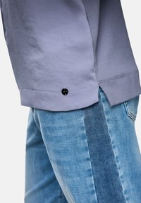 Hellblaues langärmliges Shirt mit schwarzem Logoakzent am Saum, kombiniert mit blauen Jeans, die ein horizontales Denim-Panel haben.