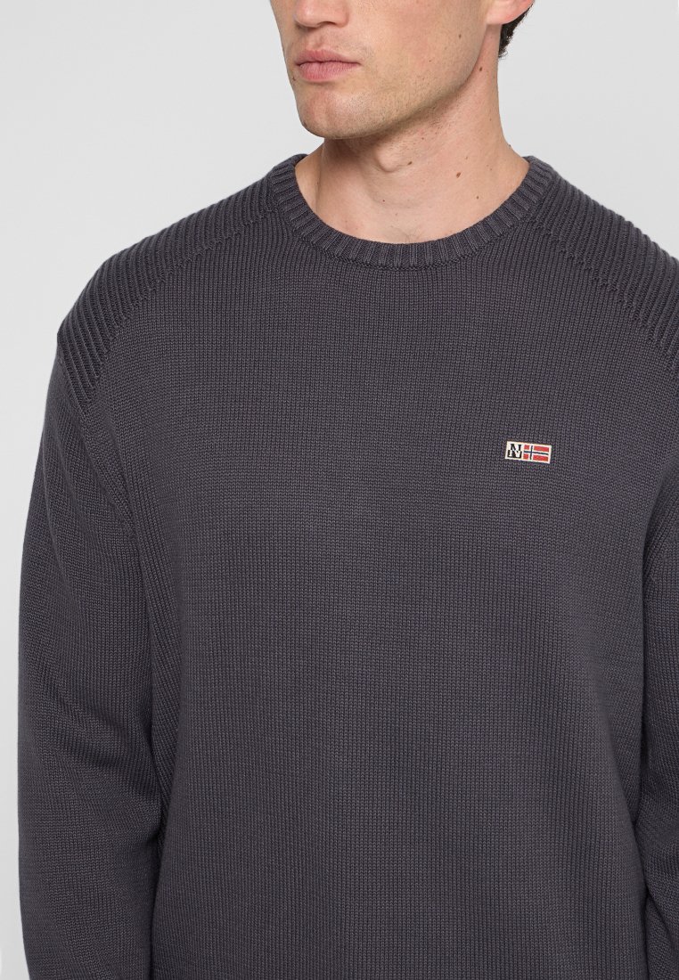 Man in een donkergrijze gebreide trui met een klein rechthoekig logo-embleem met een rood en wit vlaggetje op de borst.