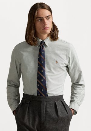 Camisa cinza-claro de botão, com colarinho, combinada com uma gravata azul-marinho com um padrão de logótipos e riscas. Calças pregueadas cinza-escuro.