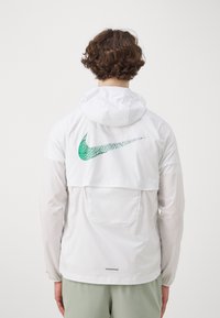 Biely kabát s kapucňou a priehľadnými rukávmi, na chrbte s zeleným a čiernym logom Nike a jednou zadnou kapsou.