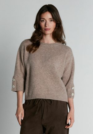 Femme portant un pull en maille beige à manches trois-quarts avec des boutons décoratifs, associé à un pantalon marron foncé à cordon de serrage.