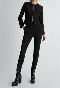 Zwarte op maat gemaakte suit met een cropped jas met een gouden rits, gecombineerd met slim-fit broeken. Draagt het met glanzende zwarte platformlaarzen.