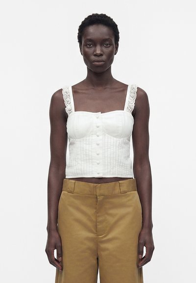 Γυναίκα που φορά λευκό crop top με κουμπιά και δαντελωτά τιράντες και παντελόνια camel με ψηλή μέση, στέκεται μπροστά σε απλό λευκό φόντο.