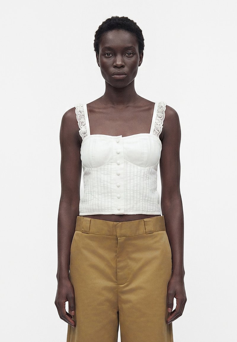 Γυναίκα που φορά λευκό crop top με κουμπιά και δαντελωτά τιράντες και παντελόνια camel με ψηλή μέση, στέκεται μπροστά σε απλό λευκό φόντο.
