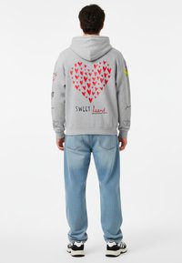 Sudadera gris con un gran gráfico de corazón rojo en la parte de atrás, texto "SWEET heart" y varios garabatos. Combinada con jeans azul claro y zapatillas deportivas.