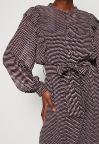 Bedrucktes Kleid mit langen Ärmeln, Rüschendetails und einem Taillengürtel. Verfügt über ein geometrisches Muster in Burgunderrot und Weiß. Hergestellt aus durchsichtiger Stoff.