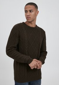 Pulover tricotat de culoare verde închis, cu un model în diamant și cabluri, având manșete și tiv din țesătură cu nervuri, care oferă textură și complexitate designului.