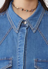 Camicia in denim con colletto classico, realizzata in tessuto azzurro chiaro e cuciture arancioni a contrasto; un choker dorato decorato con pietre colorate.
