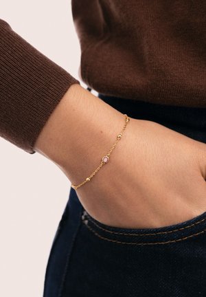 Pulsera de cadena de oro con pequeños adornos redondos y una piedra rosa central, llevada en una mano que descansa cerca de un suéter marrón y jeans oscuros.