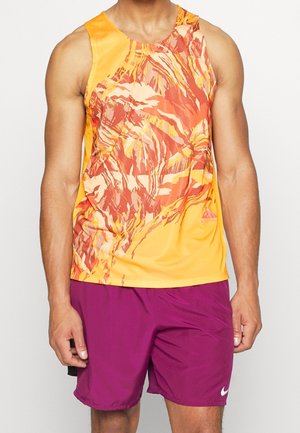 Man wearing sleeveless orange and red patterned athletic top with solid purple Nike shorts, standing against a plain background.