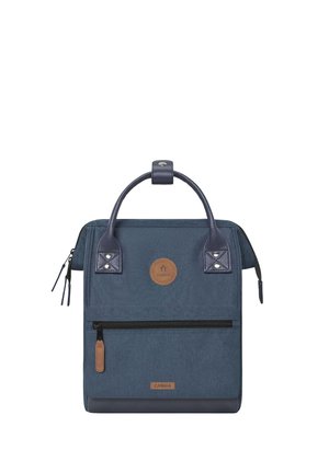 Quadratischer marineblauer Rucksack mit Ledergriffen, vorderer Reißverschlusstasche und rundem braunem Logo-Patch in der Mitte.