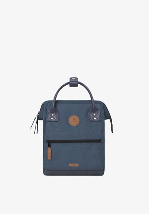 Quadratischer marineblauer Rucksack mit Ledergriffen, vorderer Reißverschlusstasche und rundem braunem Logo-Patch in der Mitte.