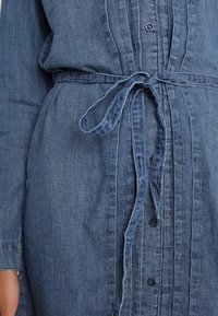 Denimskjorta i mellanblått, med framknappar, ett bälte med knytning i midjan och veckad detalj längs mitten. Texturerat tyg med långa ärmar.