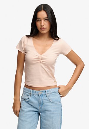 Ung kvinde med langt sort hår iført lyserød kortærmet bluse med V-hals og rynker og lyseblå jeans, poserer med den ene hånd i lommen.