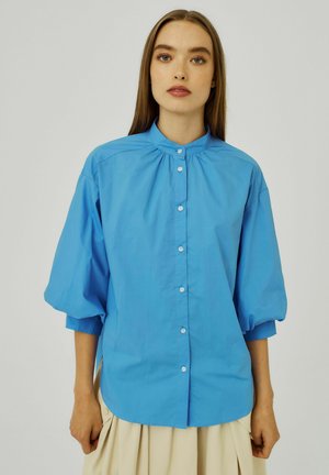 Lichtblauw, losvallend shirt met een staande kraag, poffende mouwen en knoopsluiting. Gemaakt van een gladde stof met een zachte textuur.