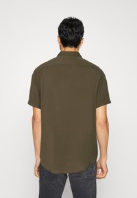 Chemise à manches courtes, couleur olive, fabriquée dans un matériau lisse avec un léger éclat. Elle possède un col classique et un ourlet arrondi.