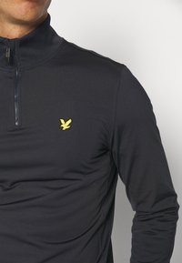 Marinblå tröja med hög krage, med en fjärdedels dragkedja och en liten gul örnlogga på bröstet. Slät, elastisk tyg.