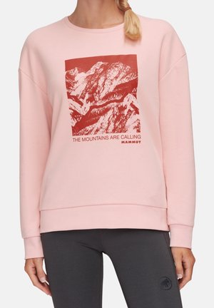 Persoon met een lichtroze sweatshirt met een rode bergafbeelding en de tekst "The mountains are calling Mammut" op de voorkant.