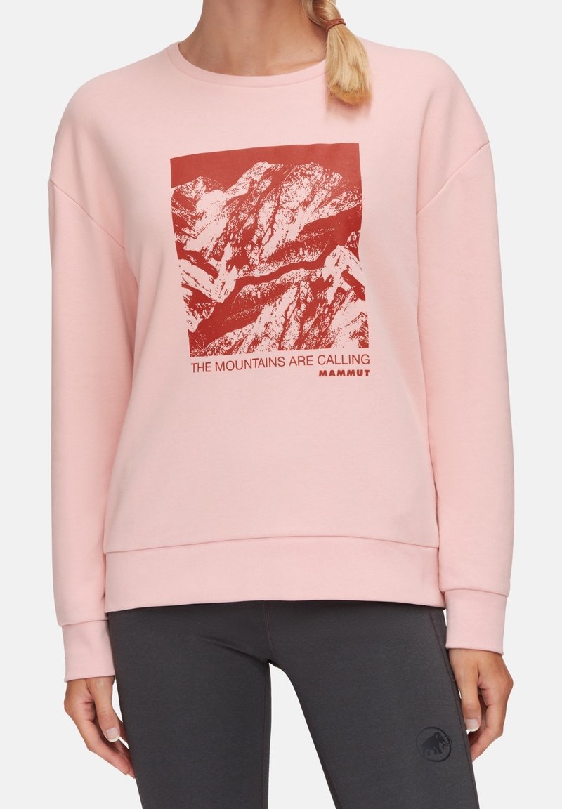 Personne portant un sweat-shirt rose clair avec un dessin de montagne rouge et le texte "The mountains are calling Mammut" sur le devant.