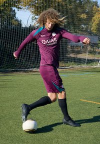 Maglia da calcio viola con accenti turchesi e pantaloni neri, abbinati a scarpini da calcio neri, mentre il giocatore calcia un pallone da calcio bianco e nero.